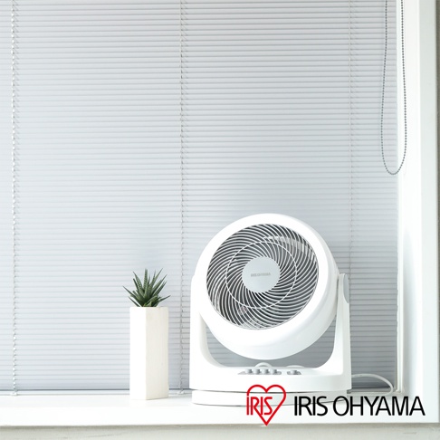 白色IRIS OHYAMA PCF-HM23 空氣循環扇，帶有可調節傾斜角度的底座，扇葉網罩上有品牌標誌。旁邊有一個白色長方形花盆，種植著綠色多肉植物。背景是淺灰色的百葉窗。