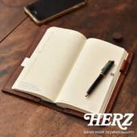 職人手作皮革旅行記事本書套，焦糖棕色，內含一本打開的日誌，一本綠色封面的筆記本，一支綠色與金色筆夾的鋼筆，以及一部黑色手機。