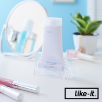 透明壓克力製的手動轉轉擠牙膏夾，夾著一管白色牙膏，背景有鏡子、植物和化妝品。