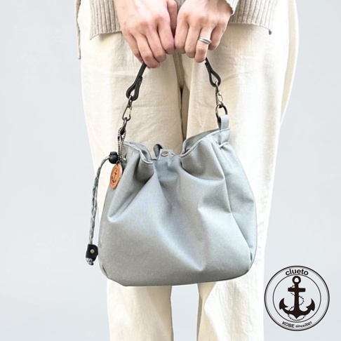 一位女性手持神戶clueto品牌的灰色防潑水多用途束口袋，包身呈現柔軟的圓弧造型，配有黑色提把及帶有船錨標誌的木質掛飾。
