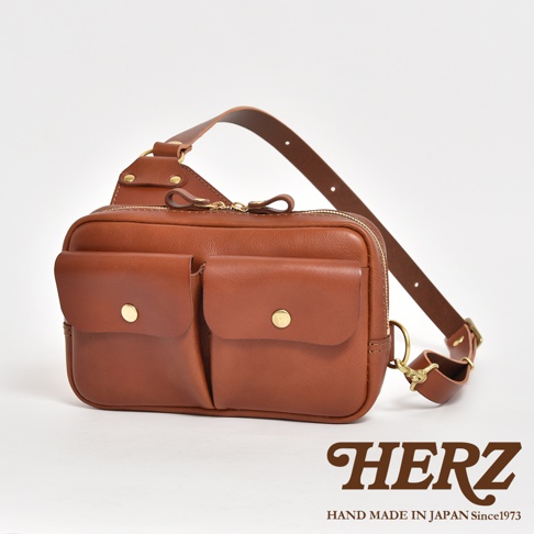 棕色皮革雙口袋斜背包，正面有兩個帶有金色釦子的翻蓋口袋，上方有拉鍊開口，搭配皮革肩帶。