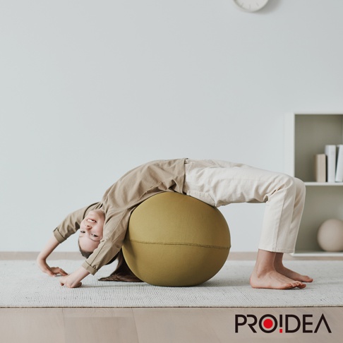 一名兒童正俯臥在一個橄欖綠色的彈力平衡球上，雙手撐地，身體向後彎曲。平衡球由針織材質製成，整體呈圓形，帶有縫線。