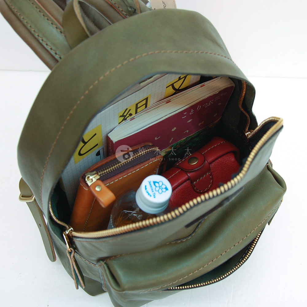 職人手作綠色皮革後背包，內部裝有筆記本、錢包、紅色皮革小包及一瓶水。