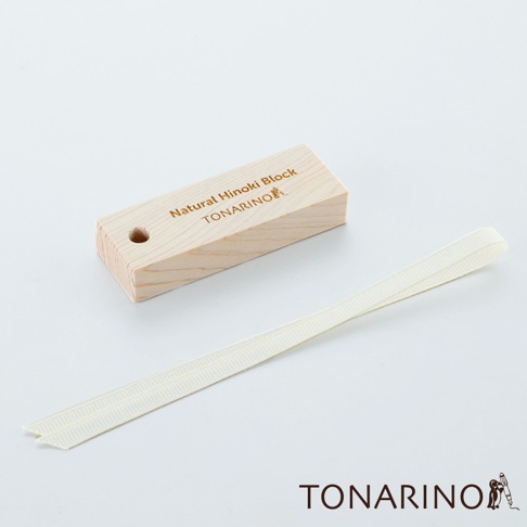 淺色木紋檜木塊，表面蝕刻有「Natural Hinoki Block TONARINO」字樣及圖示，一旁擺放著一條米白色織帶。