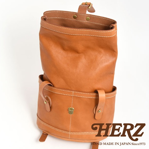 職人手作皮革雙口袋折蓋後背包，呈現自然棕色皮革，帶有雙前口袋及金屬扣環，車線明顯，風格簡約。
