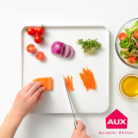 白色防溢大砧板上擺放著切片番茄、紫洋蔥、豆芽菜和胡蘿蔔絲。一只手正用刀切胡蘿蔔，旁邊還有切好的胡蘿蔔絲。右側有沙拉碗和裝有橄欖油的小碗。