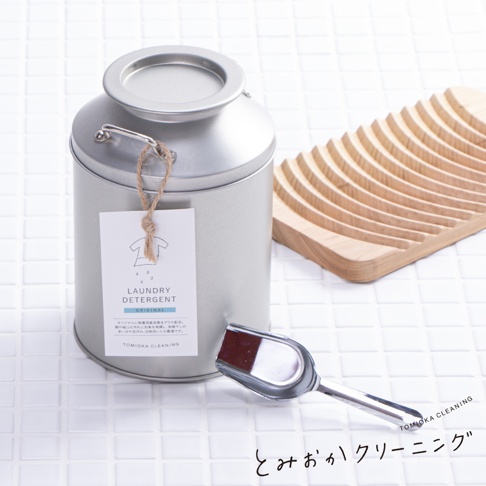 銀色牛奶罐狀洗衣粉罐，附有吊牌標示「LAUNDRY DETERGENT ORIGINAL」。一旁有木製洗衣板和銀色金屬勺子，勺子內裝有深棕色粉末。