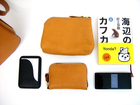 職人手作皮革翻蓋直式單肩包 (CP-96) 展示了兩個棕褐色皮革零錢包，一個黑色卡夾，一本黃色封面日文書籍，以及一部黑色摺疊手機。
