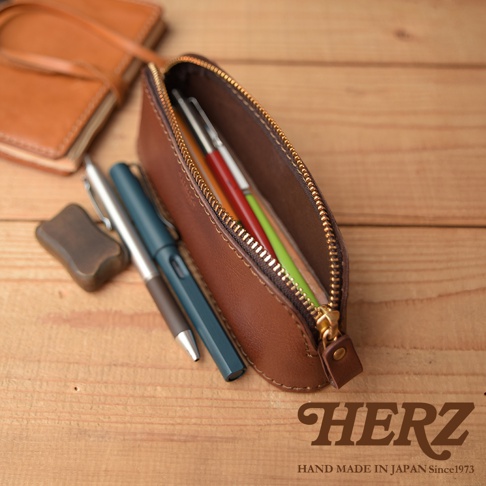 職人手作皮革三角弧形收納筆袋，棕色皮革材質，拉鍊開啟，內有數支不同顏色的筆，旁邊另有兩支筆與一塊方形皮革小物，背景為木紋桌面。