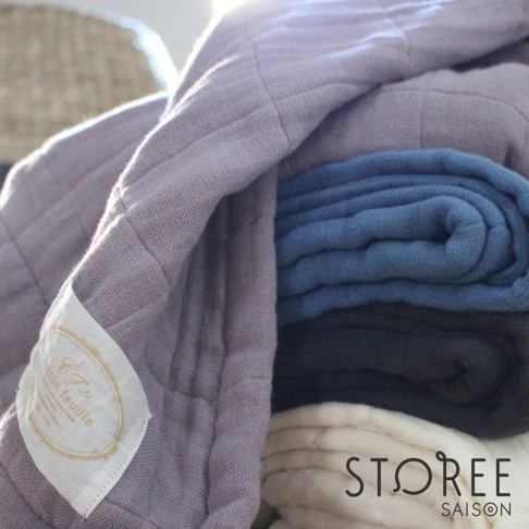 疊放的九層紗單人毯，顏色由淺至深包含淡紫色、藍色、炭灰色及米白色。布料表面有細緻的格紋縫線，左側有一枚印有品牌標誌的織標。