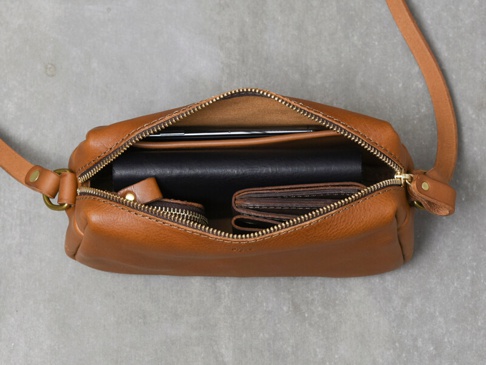 棕色皮革製成的拉鍊式單肩包，內部裝有手機、筆記本和錢包。