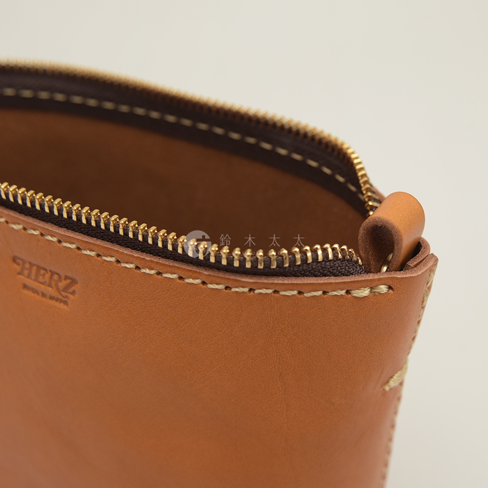 職人手作皮革三角收納包，採用淺棕色皮革，配有金色拉鍊和縫線細節。包身印有「HERZ」品牌標誌。