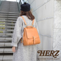 一位身穿灰色外套的女性背著一個棕褐色的皮革後背包，背包設計簡潔，有翻蓋和金屬扣環，散發著溫暖的復古氣息。