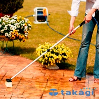 一隻手拿著帶有白色刷毛的銀色伸縮桿，連接到藍色水管，正在清潔磚砌的露台。背景有花卉盆栽和灑水器。