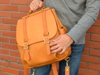 一個棕褐色皮革後背包，帶有兩個金屬扣環和皮帶，頂部有手柄，由一個人拿著，背景是紅磚牆。