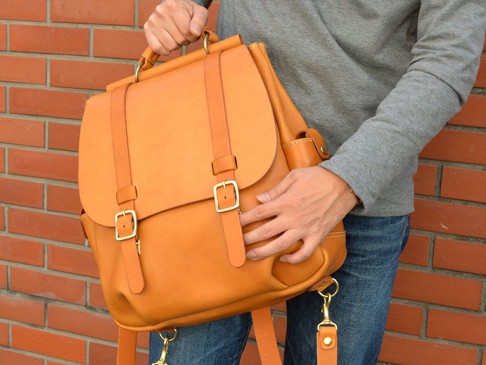 一個棕褐色皮革後背包，帶有兩個金屬扣環和皮帶，頂部有手柄，由一個人拿著，背景是紅磚牆。
