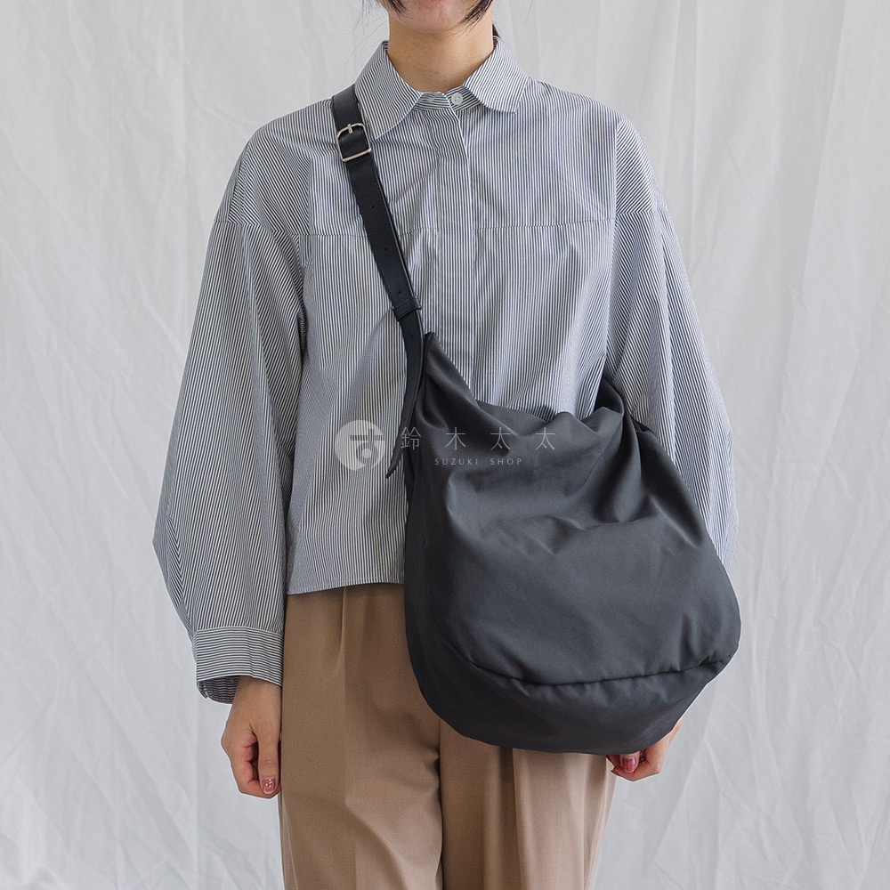 一位女子身穿藍白條紋襯衫，肩背一個黑色托特包，手持包包，身後為白色背景。