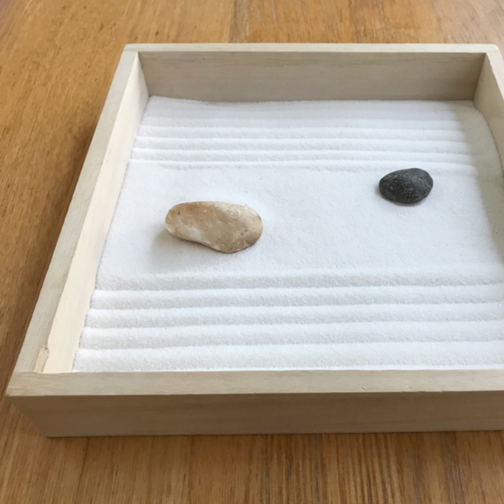 淺色木製盆中，白色細砂被耙出連續的波紋，砂上放置著兩顆大小、顏色各異的石頭，一顆為淺棕色鵝卵石，另一顆為深灰色小石。