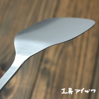 不鏽鋼萬用鍋鏟，呈銀灰色，寬扁的鏟面帶有弧度，邊緣光滑，手柄為長條形，整體材質為金屬，背景為木紋紋理。