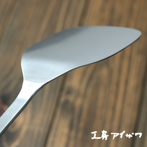 不鏽鋼萬用鍋鏟，呈銀灰色，寬扁的鏟面帶有弧度，邊緣光滑，手柄為長條形，整體材質為金屬，背景為木紋紋理。