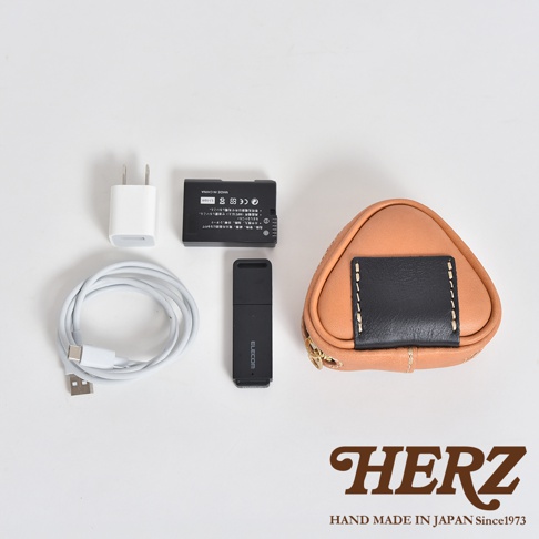 一個棕色皮革製成的三角飯糰造型小物收納包，搭配黑色皮革裝飾和縫線，以及金色拉鍊。一旁有白色充電器、黑色電池、白色充電線和黑色USB裝置。