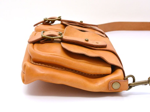 職人手作皮革雙口袋肩背包，天然皮革材質，色澤為淺棕色，配有黃銅色金屬扣環和鉚釘，展現簡約復古風格。