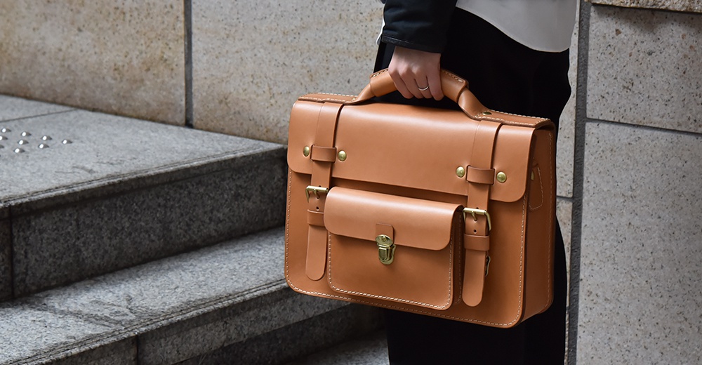職人手作學院風兩用公事包，採用 tan 色皮革製成，配有黃銅色金屬扣環和可調節的側邊皮帶。包款正面有兩個翻蓋式口袋，上方有提把，整體呈現復古學院風格。