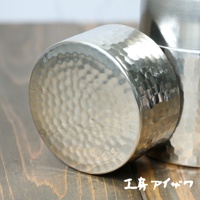 銀色不鏽鋼錘目紋茶葉罐，表面帶有不規則的錘打紋路，光澤感強，搭配木質桌面背景。