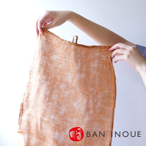 一雙手拿著橙色三層蚊帳棉織廚房拭巾，拭巾質地輕薄，有細微的白色斑駁紋理，頂部邊緣有金色編織掛環。