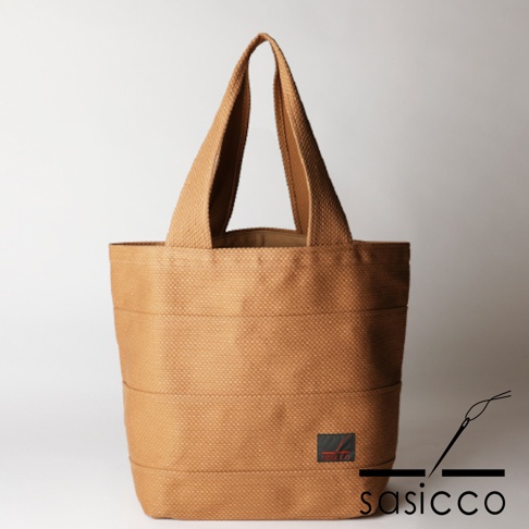 三河木綿刺子織大容量托特包，採用棕褐色棉質刺子織布料製成，具有多層水平縫線設計，包身寬大，配有兩條提把，底部右側有黑色小標籤。