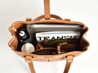 職人手作皮革原子釦托特包，淺棕色天然皮革製成，包內裝有文件夾、雜誌、錢包、摺疊傘和白色圓形物品，展現包款的收納空間與質感。