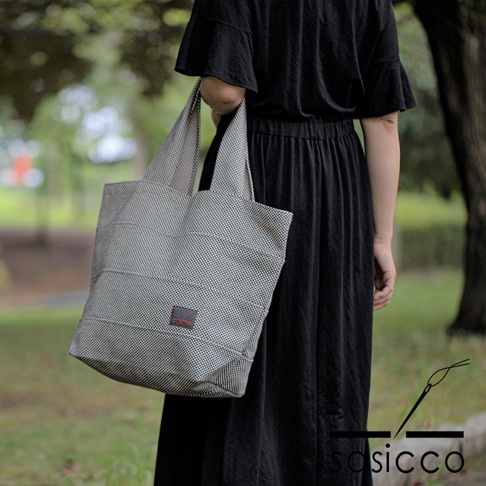 一位女性手提一個灰色帶有黑點刺子織紋路的帆布托特包，包身有水平縫線，左側有紅色品牌標籤。