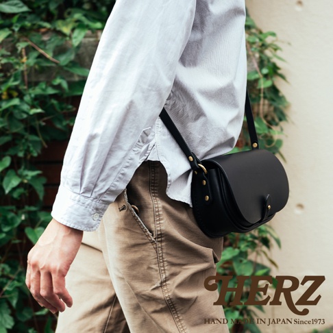職人手作黑色皮革迷你馬鞍包，設有可調節肩帶，以黃銅金屬扣固定，包身線條圓潤，展現精緻工藝。