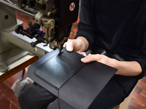 職人正在使用工業縫紉機縫製黑色皮革翻蓋迷你單肩包。包袋採用光滑皮革製成，邊緣有精細的縫線。