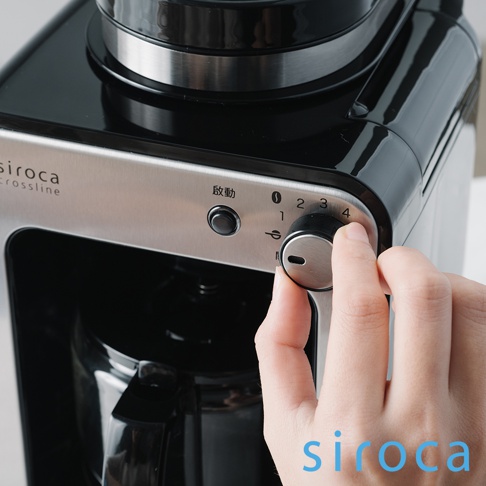 SC-A1210 自動研磨咖啡機，黑色機身搭配不鏽鋼前面板，上面有「啟動」按鈕和刻度旋鈕，使用者正用手指調整旋鈕。