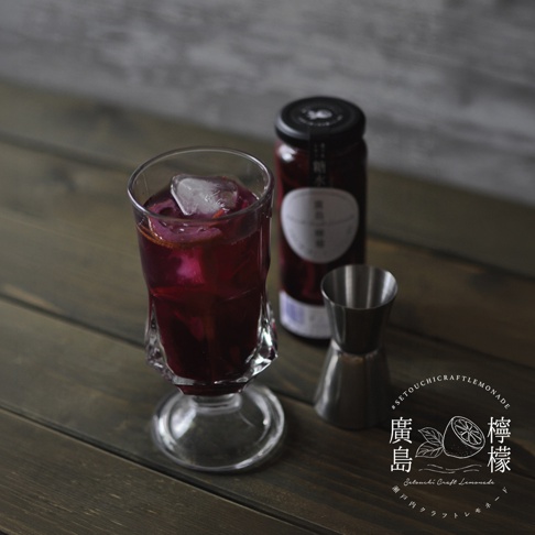 深色木質餐桌上放置著一杯深紫色的藍莓檸檬釀飲品，杯中裝滿冰塊與果片，後方有一瓶同款果釀與一個不鏽鋼量杯，畫面右下角帶有廣島檸檬的手繪標誌與日文標籤。