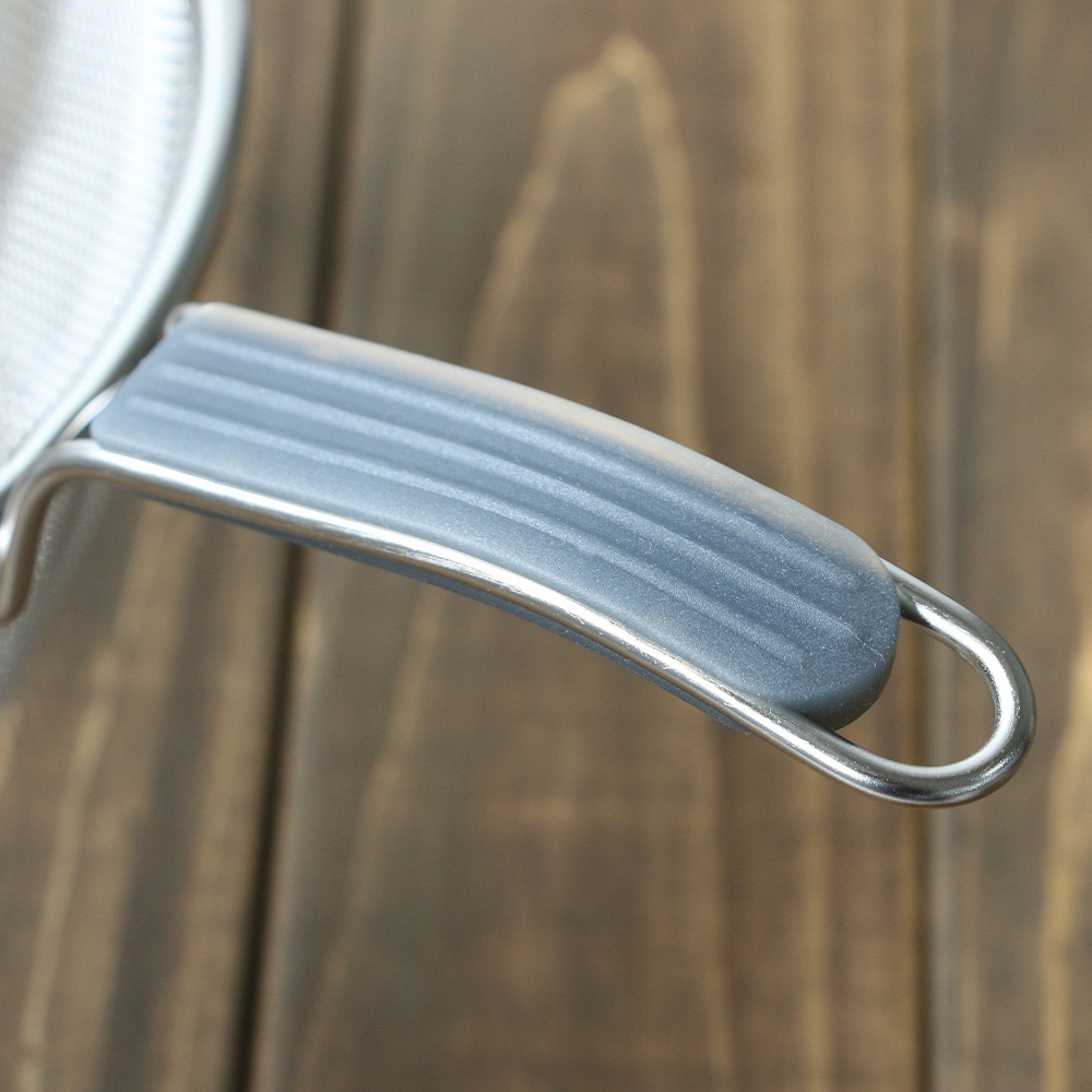 不鏽鋼濾網的銀色金屬框架，搭配一個灰色矽膠防滑握柄，握柄上有橫向紋路，整體呈近拍特寫。