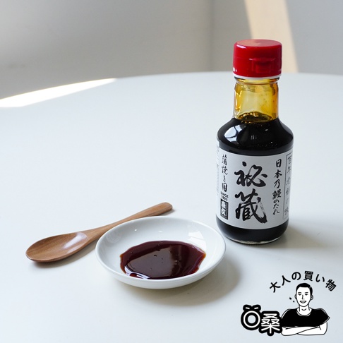 玻璃瓶裝深棕色秘藏燒烤鰻魚沾醬，瓶蓋為紅色，標籤上有日文文字。醬汁倒在一個白色小碟中，旁邊有一支木製湯匙。