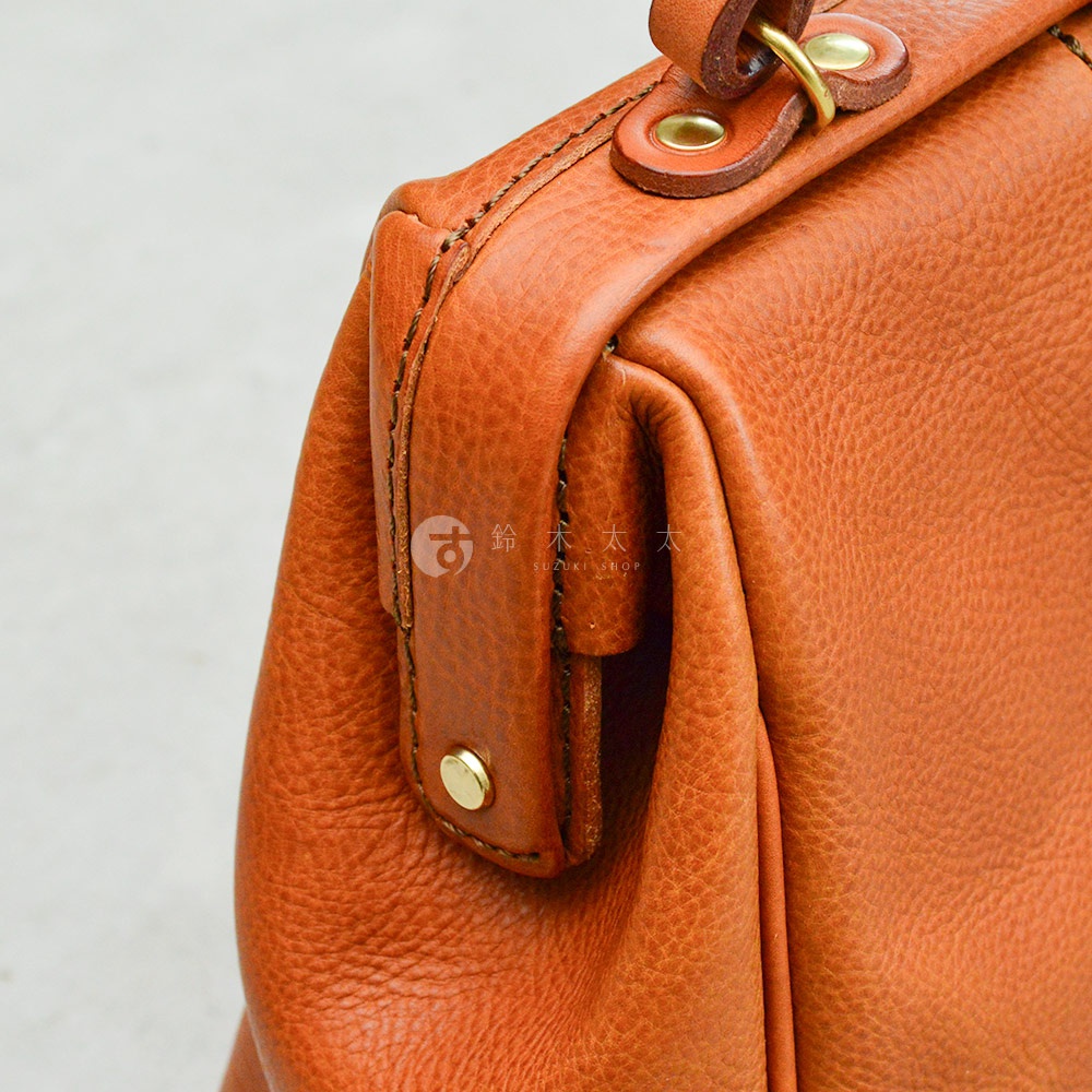 棕褐色皮革手作托特包，特寫包側邊的方形口金結構、金屬鉚釘及縫線細節，皮革表面紋理清晰。
