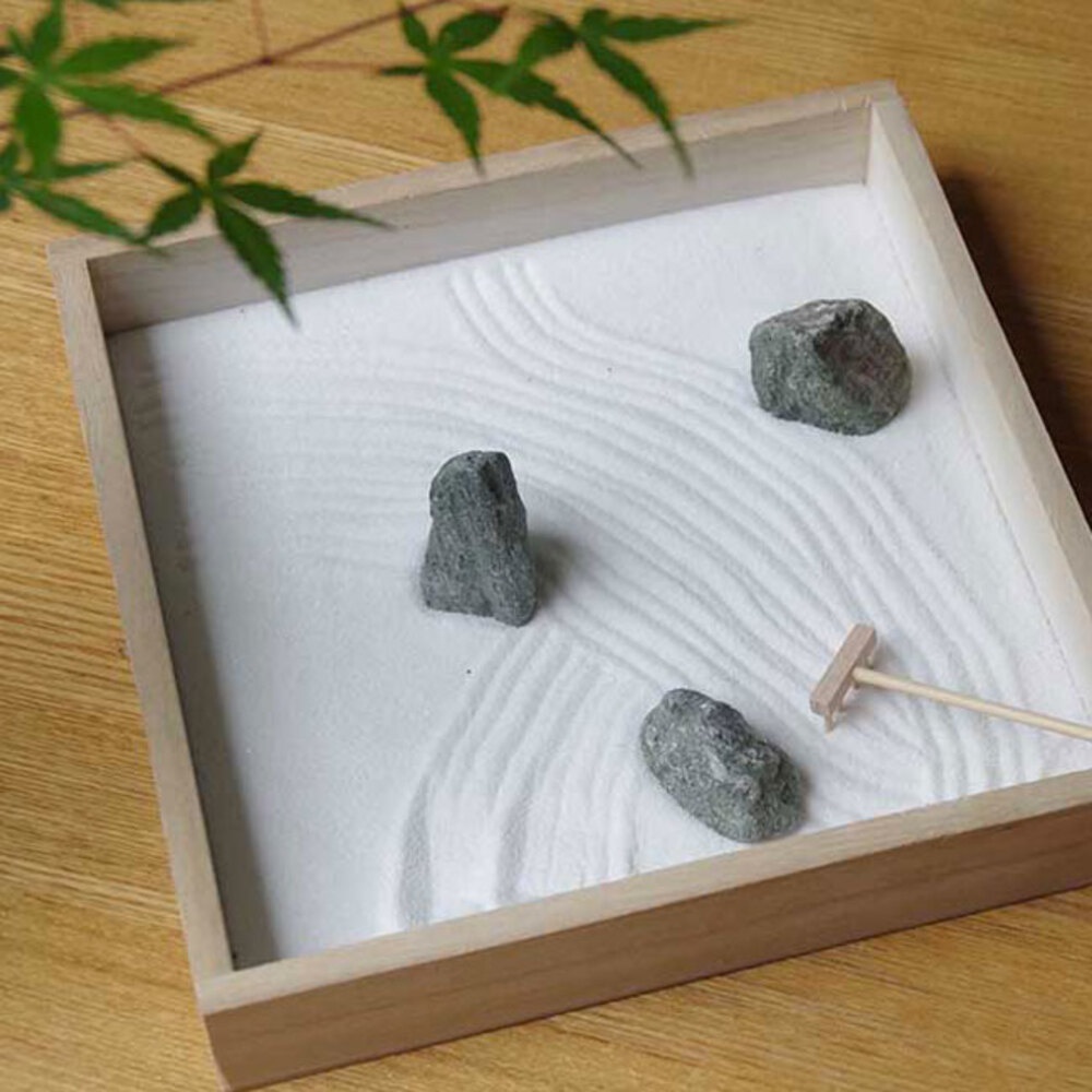 日式枯山水模型，木製淺盤盛裝白色細沙，耙出波紋，擺放三塊灰黑色不規則石塊，其中一塊旁有迷你木耙。