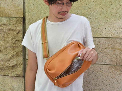 棕色皮革立體運動斜背包，配有卡其色織帶和金色金屬扣環，展示一個拉鍊隔層，內有一瓶水。