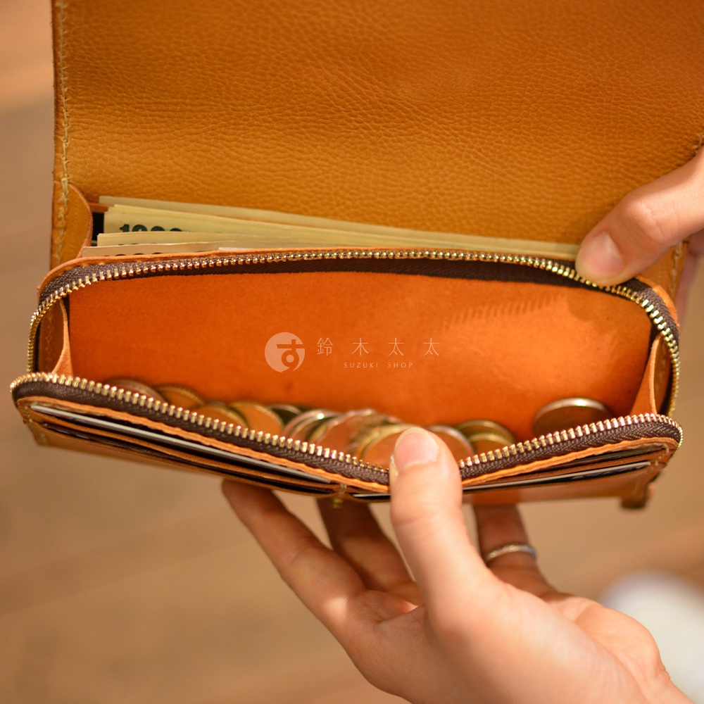 職人手作皮革長夾，外觀為質感棕褐色，內部襯裡為亮橘色，夾層可見鈔票與硬幣，拉鍊為金色金屬材質，由雙手握持展示。