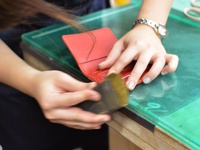 一雙手正在打磨一個紅色皮革三面雙折票券收納夾的邊緣，皮革表面光滑，縫線清晰，收納夾內有隔層。