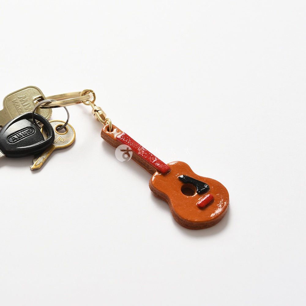 職人手作皮革吉他造型鑰匙圈，主體為棕色皮革，吉他琴頸為紅色，琴身點綴黑色與紅色細節，附有金屬鑰匙圈。