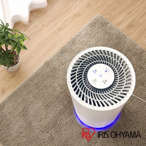 白色圓柱形空氣清淨機，頂部有放射狀的白色扇葉和藍色指示燈，底部散發著藍光，置於米色絨毛地毯上。