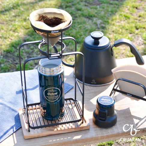 黑色金屬支架上，裝有濾紙和咖啡粉的手沖濾杯，下方置有一個藍綠色金屬保溫瓶。背景為黑色帶溫度計的細嘴咖啡壺，以及裝有濾紙的支架。