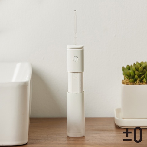 白色圓筒狀便攜沖牙機，設有透明噴嘴和圓形按鈕，機身分為三個部分，底部為半透明材質，置於木質檯面，背景為白色牆壁和盆栽。