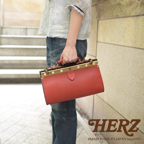 一個紅色皮革復古口金兩用手提包，搭配金色金屬配件，由手提握把提起，展示於一位穿著牛仔褲和淺色上衣的人身上。