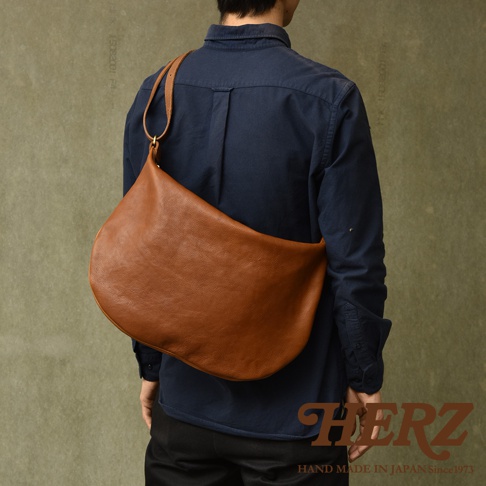 棕色皮革製成的職人手作Organ單肩半月包，斜挎於身穿深藍色襯衫的模特兒身上，包款呈彎月形狀，材質觸感紋理清晰可見。