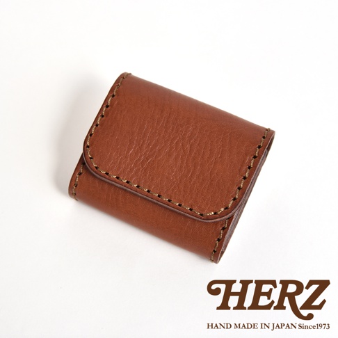 一張由日本HERZ職人手作的棕色皮革雙折零錢包，材質選用Stare皮革，表面呈現自然的紋理與色澤，並以粗縫線手工縫製。商品右下角印有HERZ品牌商標與HAND MADE IN JAPAN字樣，整體展現簡約質樸的手工質感。